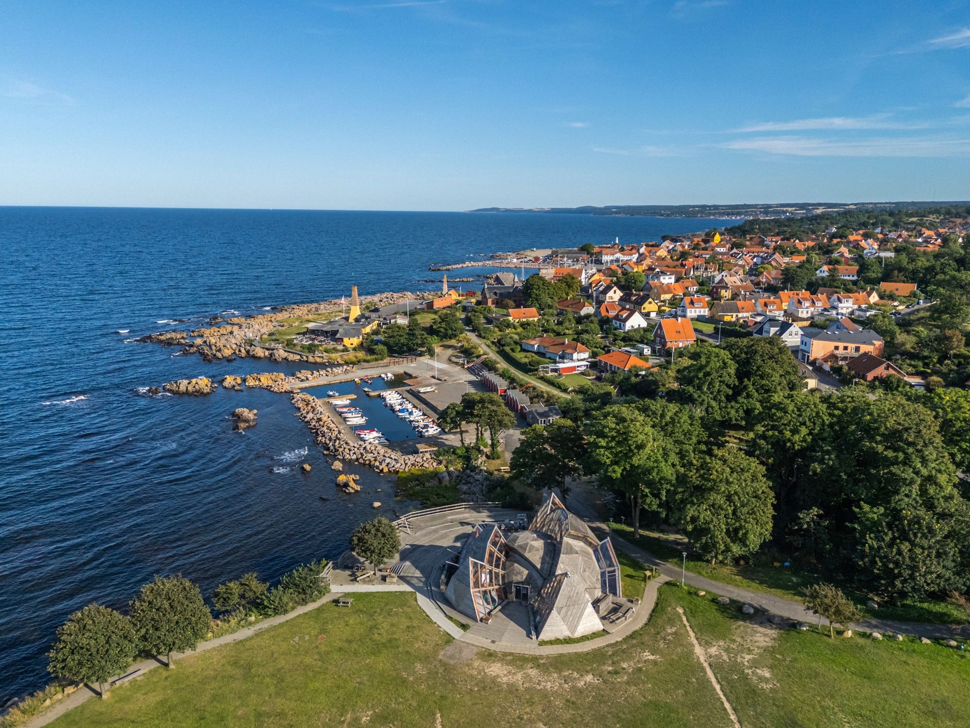 Luftbildaufnahme von Allinge mit dem kleinen Hafen, dem markanten modernen Dome-Gebaeude im Vordergrund und der Ortschaft am Ostseeufer.