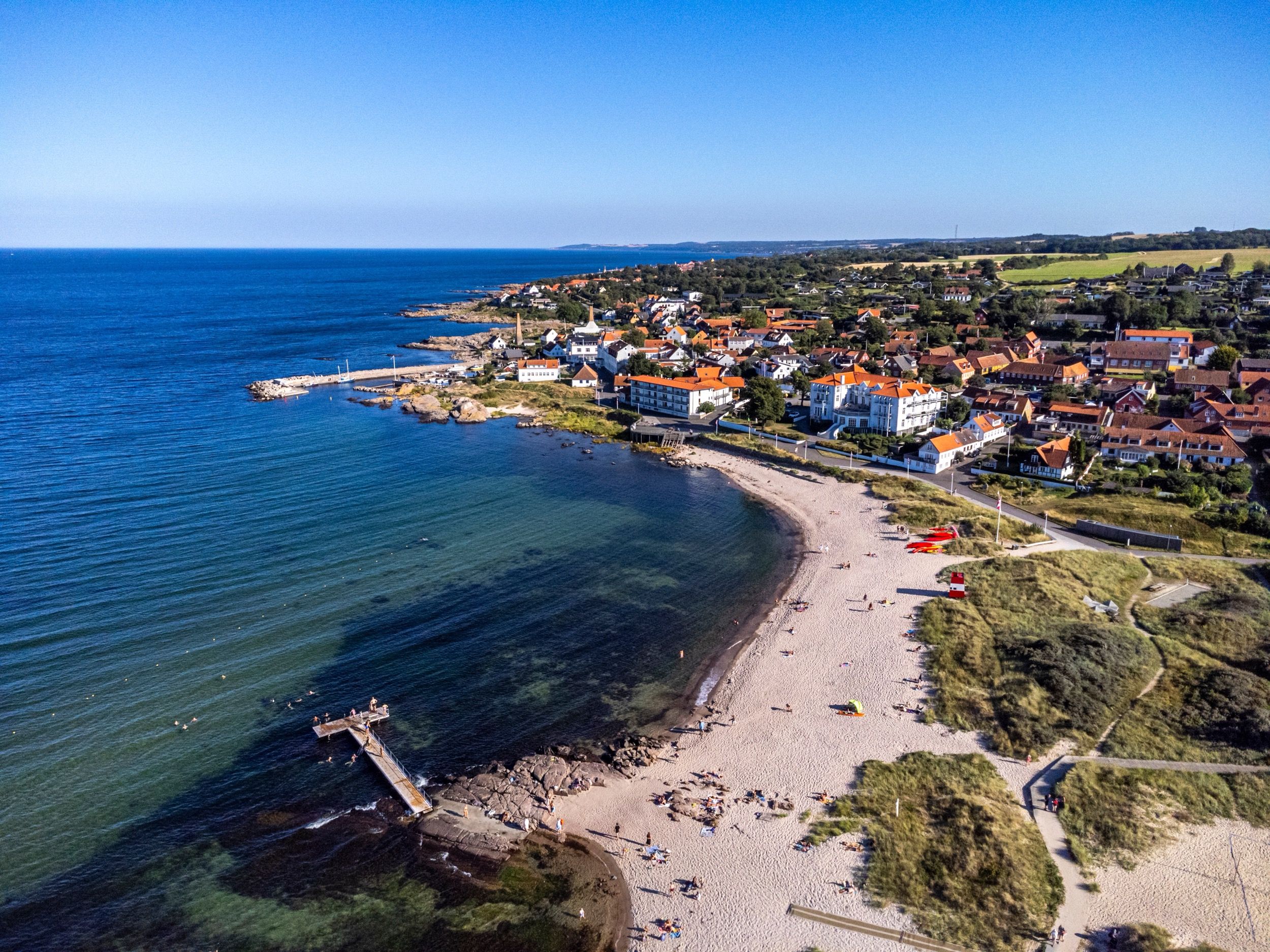Luftaufnahme des Kueroerts Sandvig mit seinem geschwungenen Sandstrand, dem kleinen Hafen und den charakteristischen weissgetuenchten Haeusern mit roten Ziegeldaechern.