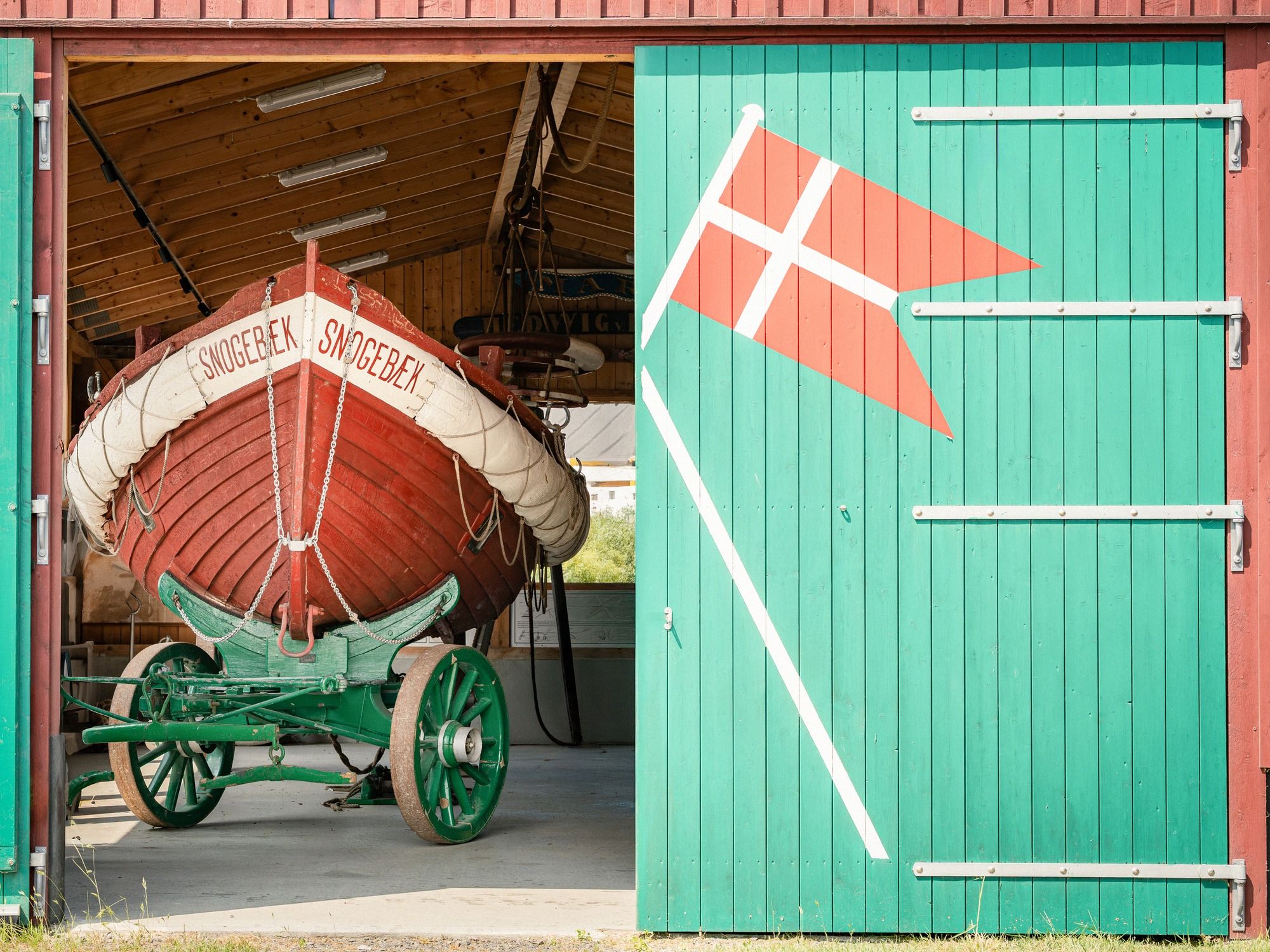 Im grünen Holzschuppen am Hafen von Snogebæk steht ein altes rotes Rettungsboot auf einem historischen Transportwagen, die Tür trägt eine aufgemalte dänische Flagge.
