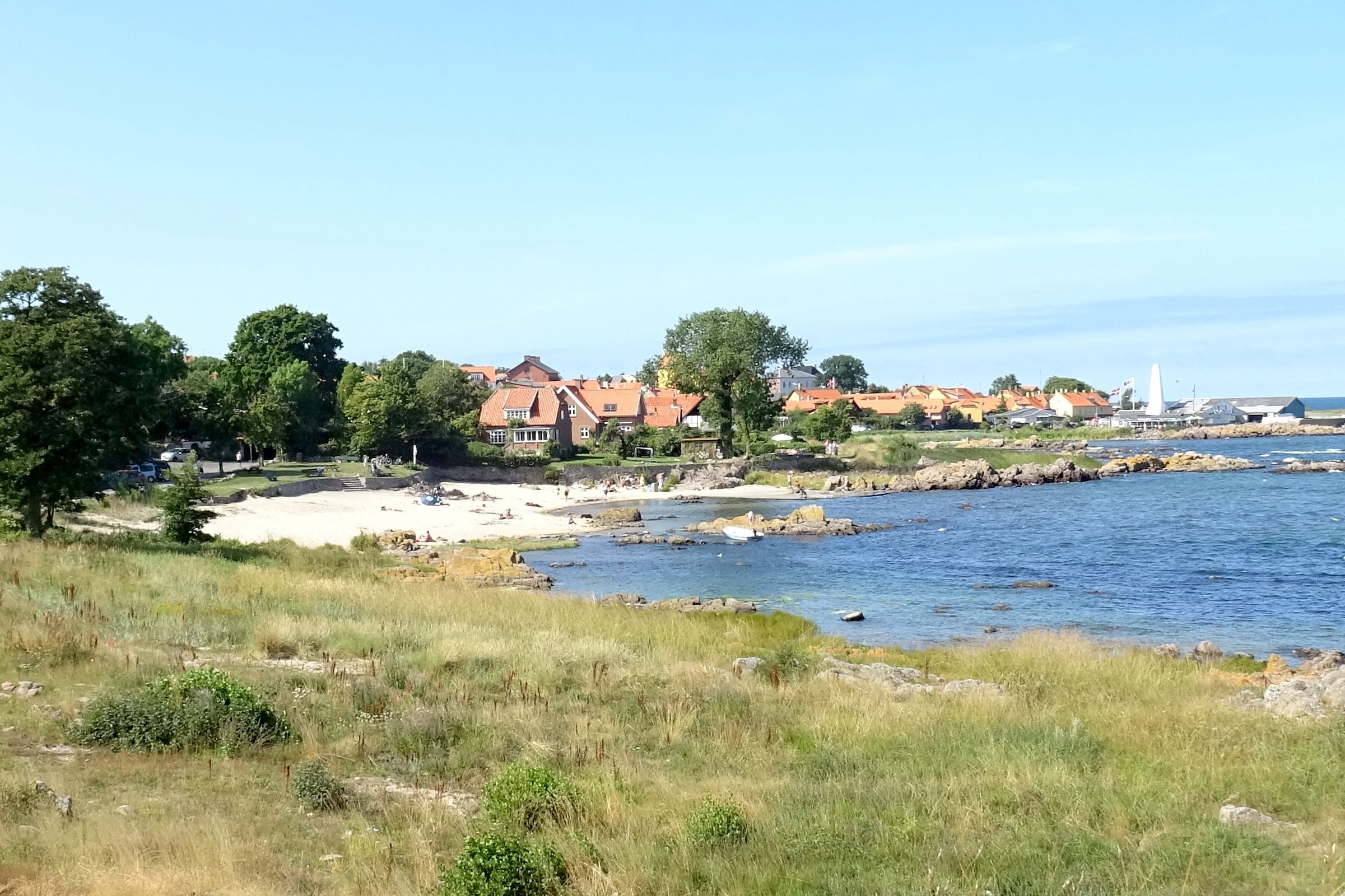Blick auf den Næs Strand bei Allinge mit kleinem Sandstrand, Felsküste und den roten Dächern der Stadt im Hintergrund.