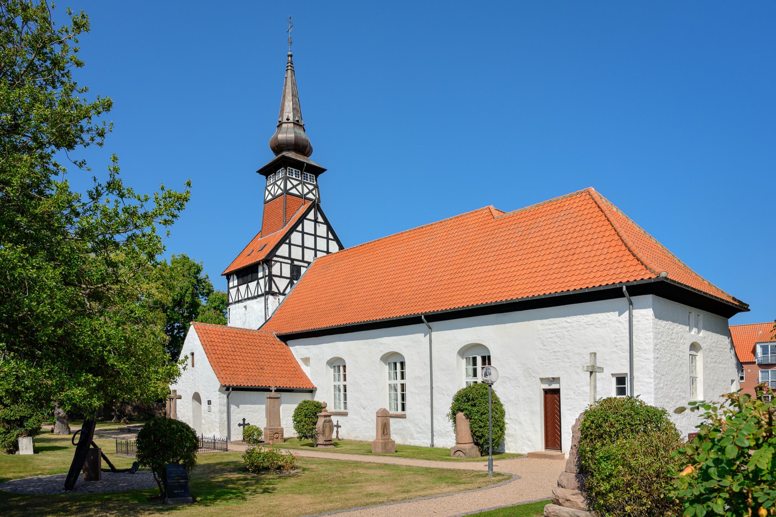 Die spaetgotische Nexø Kirche mit ihrem markanten Fachwerkturm und orangenem Ziegeldach liegt eingebettet in einen gruenen Friedhof unter blauem Himmel.