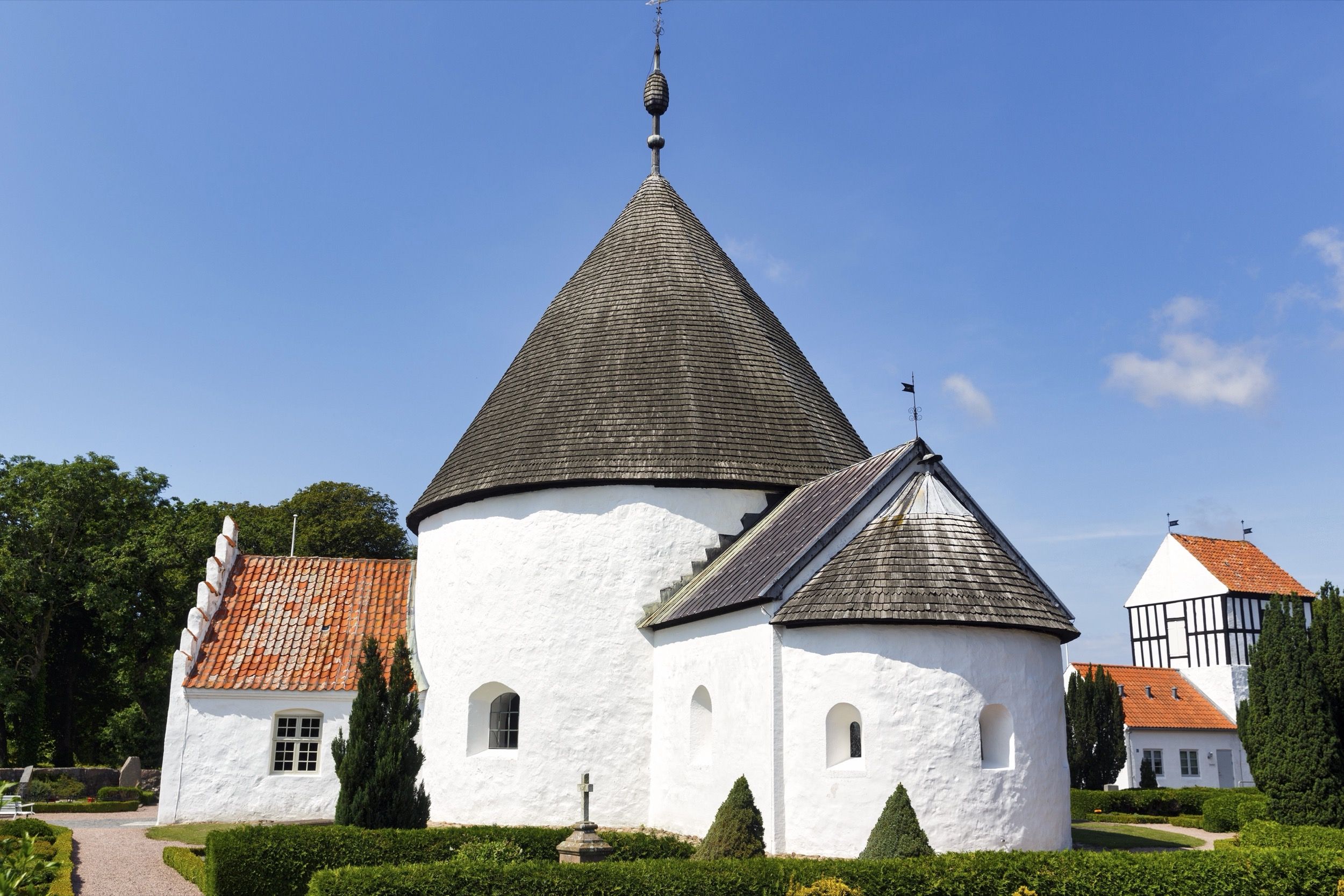 Die weiß getünchte Nyker Kirke — kleinste der vier Bornholmer Rundkirchen — mit spitzem Schieferdachhelm und gepflegtem Kirchgarten an einem sonnigen Sommertag.