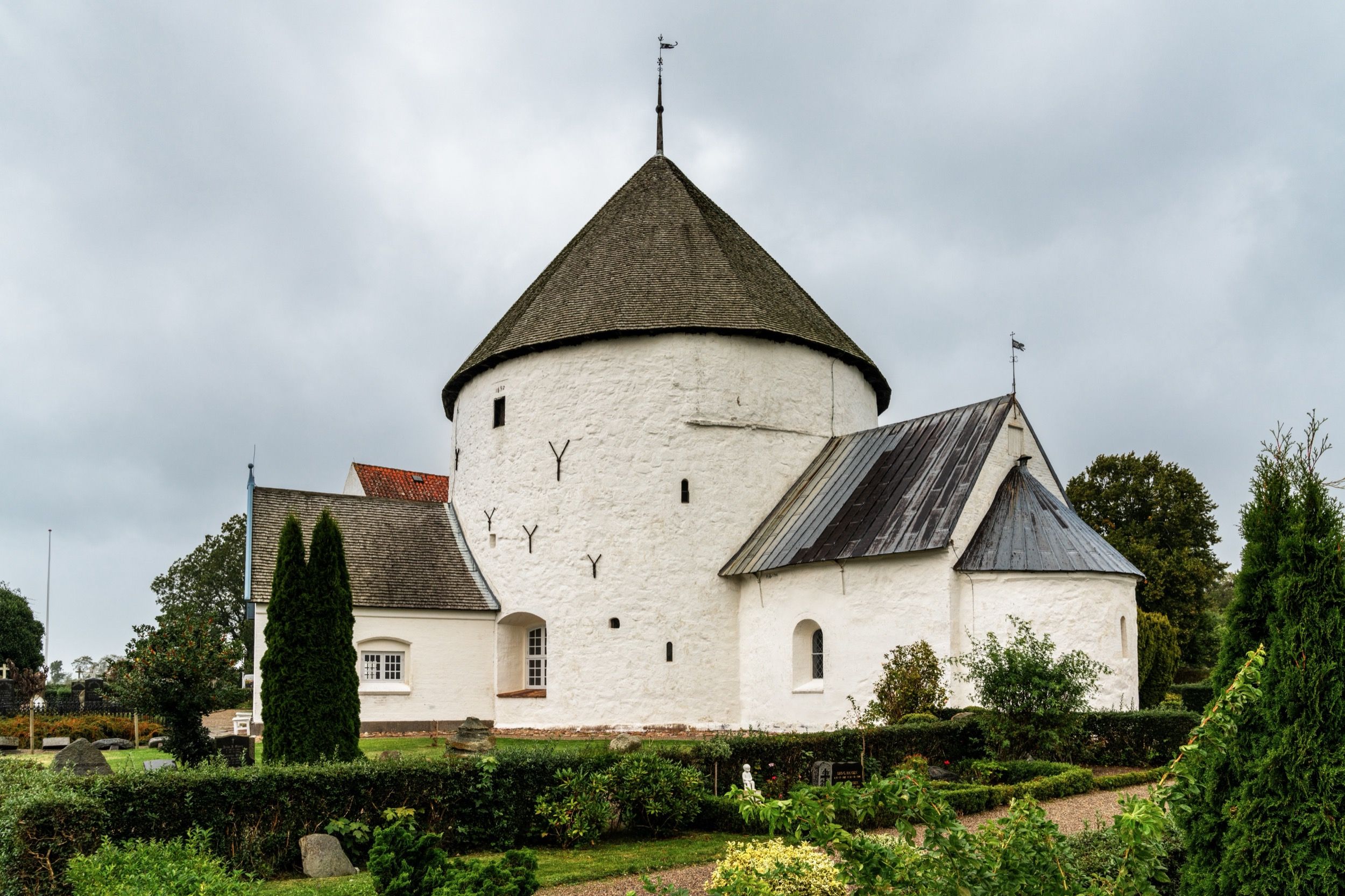 Die Nylars Kirke aus dem 12. Jahrhundert — eine der vier mittelalterlichen Rundkirchen Bornholms — umgeben von gepflegtem Friedhofsgarten unter bewölktem Herbsthimmel.