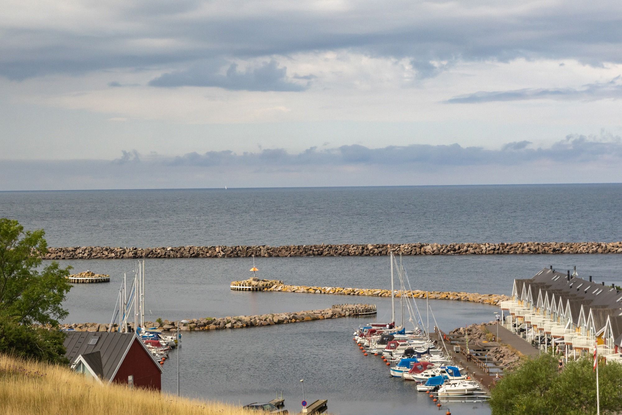 Uebersicht ueber den kleinen Hafen von Hasle mit Steinmole, Segelbooten und den weissen Bootshaeusern am Ufer unter bewolktem Sommerhimmel.