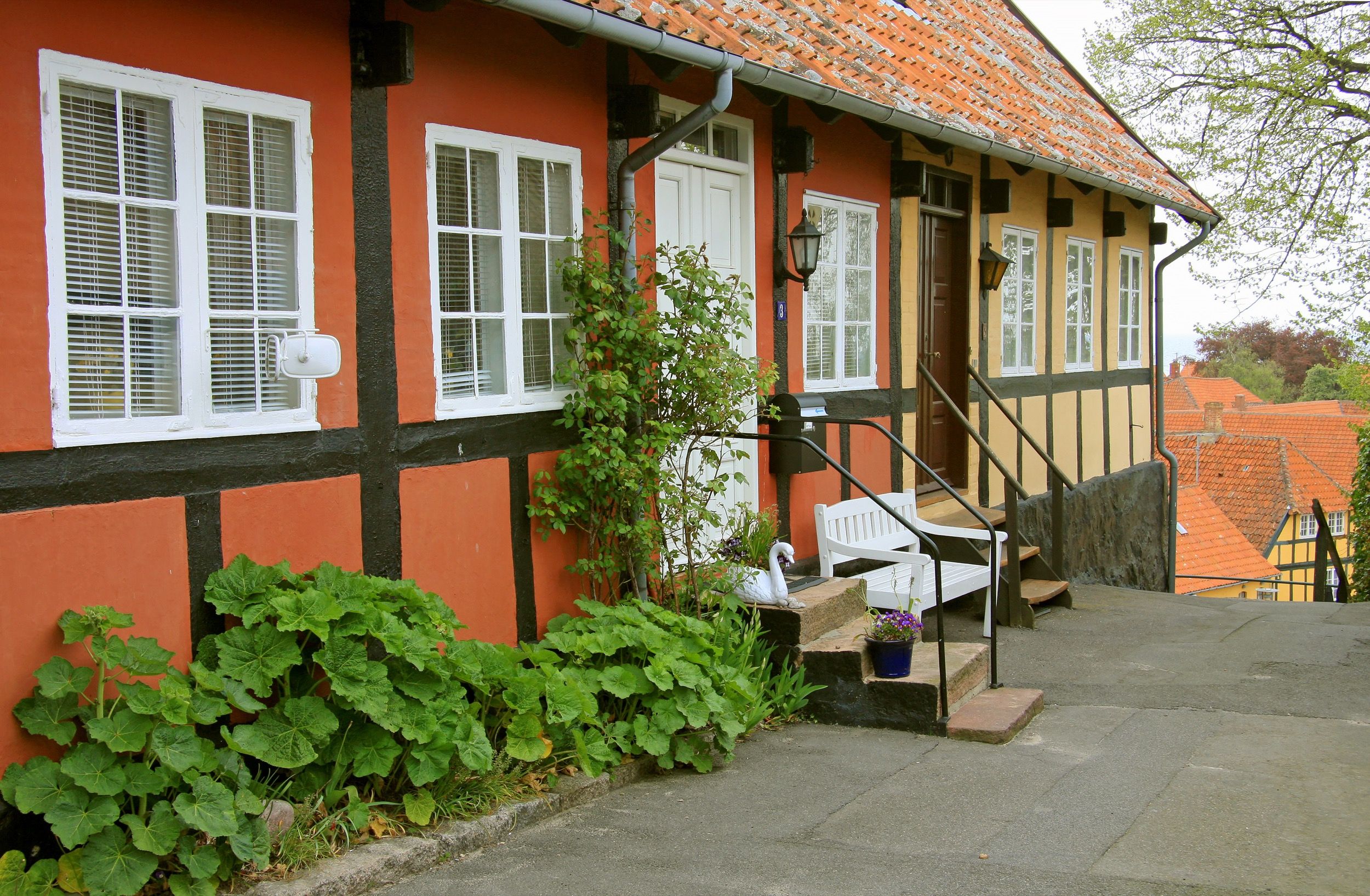 Ein rot-schwarz gestrichenes Fachwerkhaus mit weissen Fenstern und einer kleinen Treppe mit Bank praesentiert sich in einer gepflasterten Gasse von Svaneke.