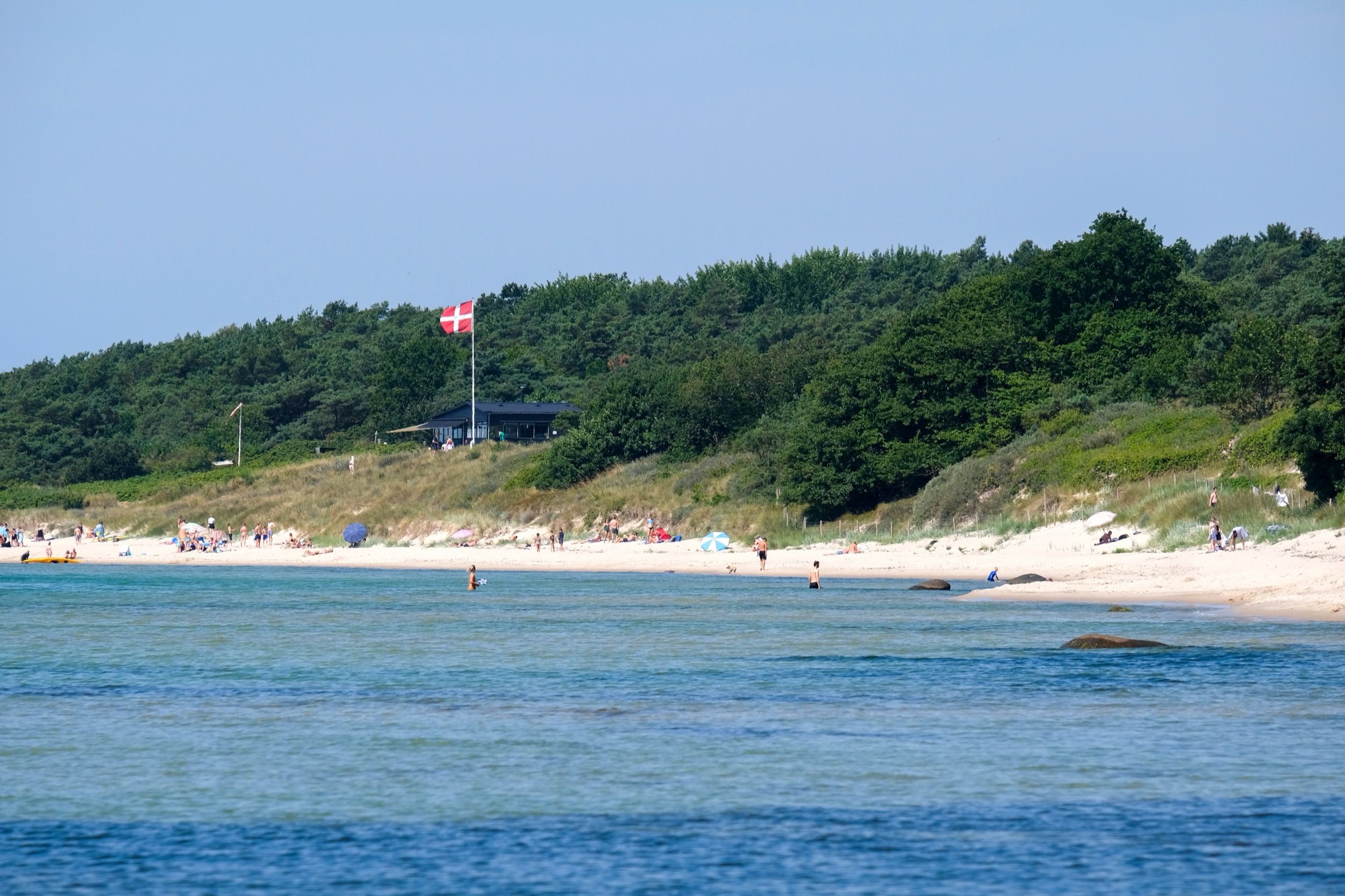 Der Sandstrand bei Sømarken an einem sonnigen Sommertag mit türkisfarbenem Wasser, Badegästen und einem Sommerhaus mit dänischer Flagge hinter der Düne.