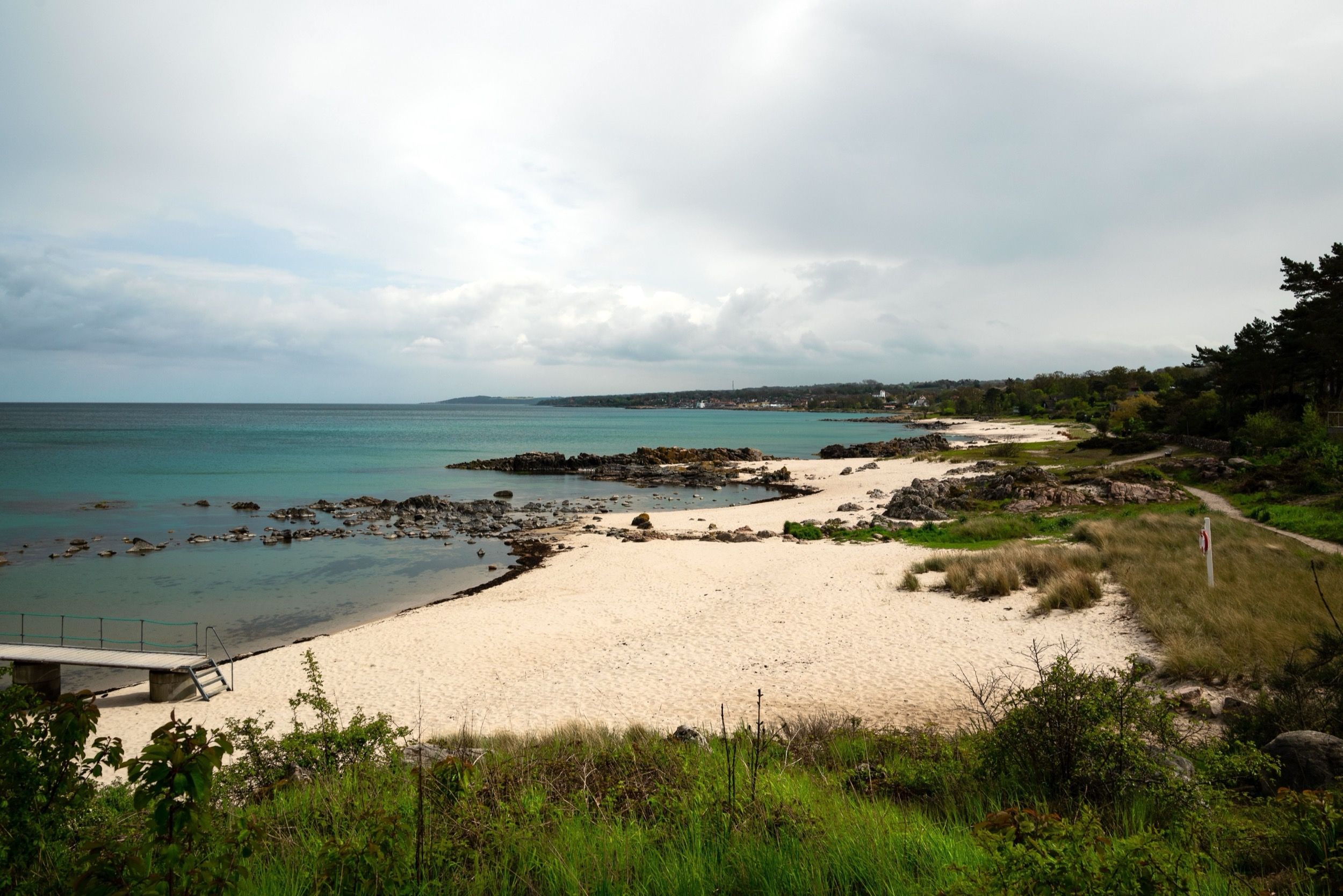 Weiter Blick auf die felsige Küstenbucht bei Tejn mit türkisfarbenem Ostseewasser, einem kleinen Sandstreifen und einem Weg entlang der bewachsenen Klippen.