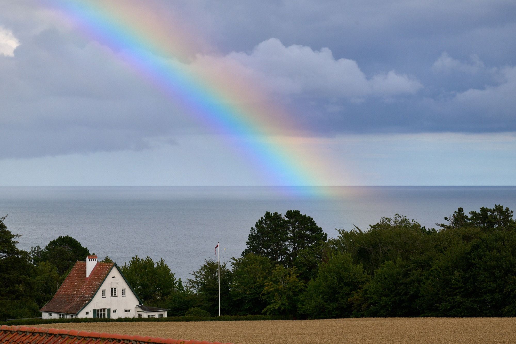 Ein leuchtender Regenbogen spannt sich über die Ostsee, im Vordergrund ein weißes dänisches Landhaus umgeben von Bäumen und einem Gerstenfeld.