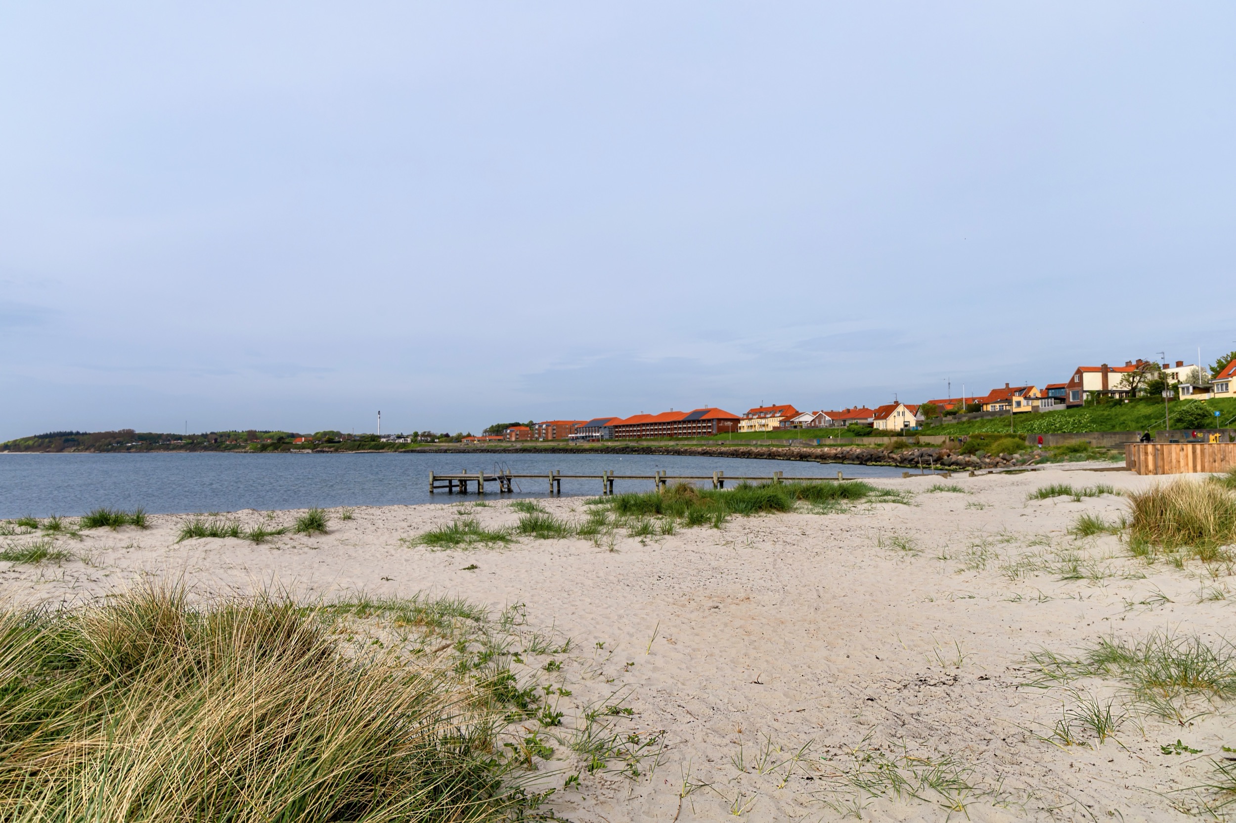Wide sandy beach with dune grass in front of Rønne's town skyline and a wooden jetty